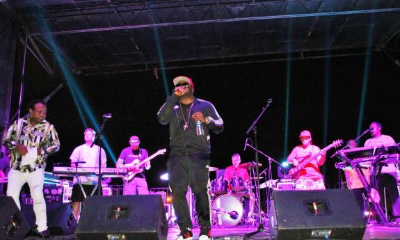 A Raggae band performs at an outdoor festival, with rose-colored lights on the stage.