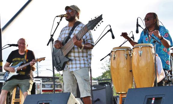 Three band members, playing guitars and conga drums perform on an outside stage during the day