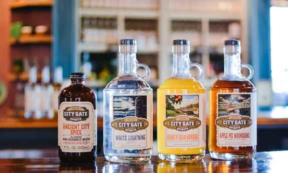 Three flavors of City Gate Sprits Moonshine and a bottle of their mixer, on a wooden counter in front of blue and white shelving