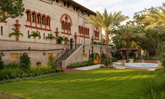 A photo of a landscaped garden and lawn, with water feature and palm trees with stairs to an historic building with red-trimmed windows