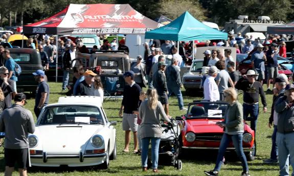 A photo of an auto show with a white and a red sport car in the foreground, people, and awning tops in the background