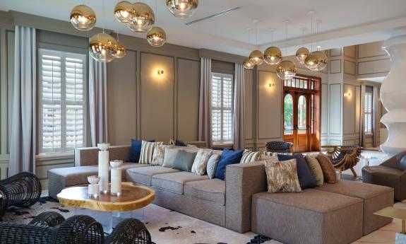 The lobby of a grand hotel, with brass globe lights, large sofas and ottomans in a warm beige and cream and wood 