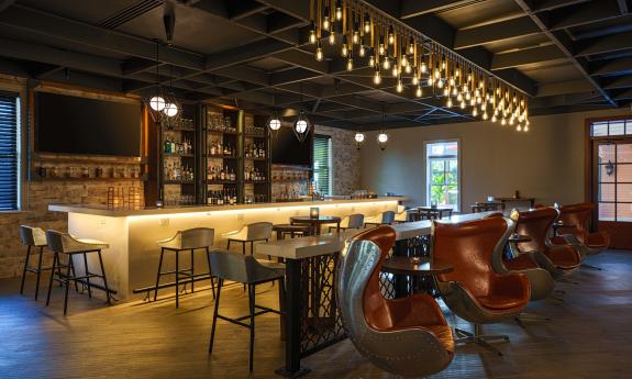 A wide shot of a bar with a cream-colored bar in the background and red chairs at bar tables in the foreground