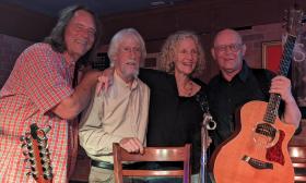 Four musicians, David Watt Beasley, Bob Patterson, Katherine Archer, and John Dickey stand together on stage with John holding a guitar