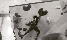 A black and white photo of a climber on an indoor climbing wall, stretching to reach the next handhold