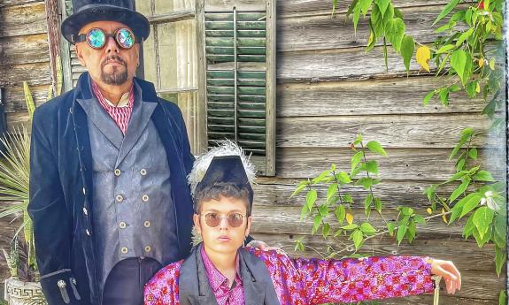 Two men, one standing and one seated, wearing steampunk garb, and next to an old wooden house, 