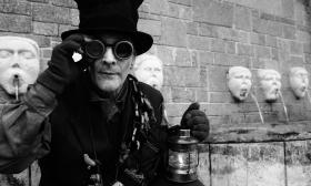 A Black and White photo of a man in steampunk garb standing near a fountain