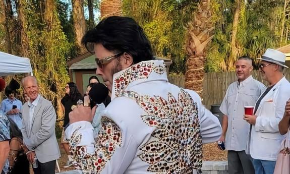 An Elvis impersonator performs outdoors, wearing a white jacket with multi-colored sequins