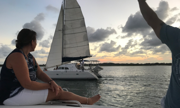 A couple on the deck of a sailboat, waving to people on a nearby sailing catamaran