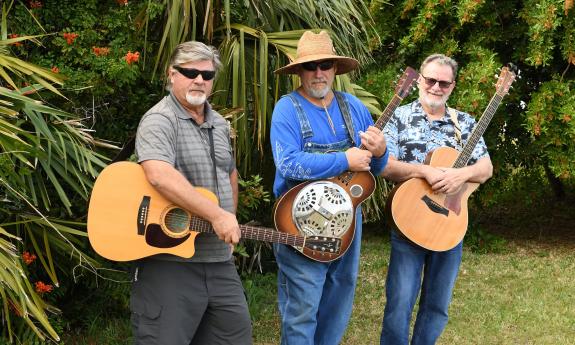 The trio Crab Creek, standing with their instruments outside among palm trees.