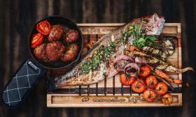 A charred board with whole fish and vegetables prepared on an Asador