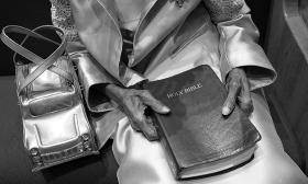An old woman's hands clutch a Bible on her well dressed lap with a trendy car shaped purse on the pew to her left