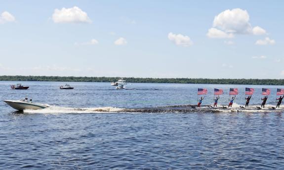 A boat towing a line of water-skiers, all holding American flags, on a lake in Florida