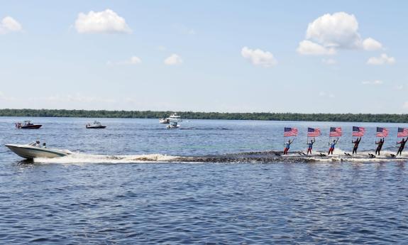 A boat towing a line of water-skiers, all holding American flags, on a lake in Florida