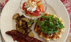 A plate of breakfast with strawberries and cream, a waffle, bacon, and home fries on a white plate with red tablecloth