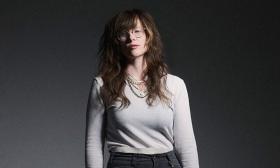 Americana Musician S.G. Goodman posing stoically in front of a gradient grey background. She is a white woman with long hair wearing necklaces and a pale top