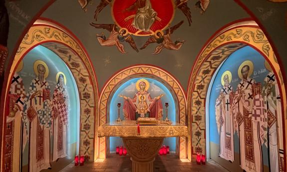 A marble altar with a gold cloth is surrounded in Byzantine-style murals of saviors, angels, and saints. Gold, red, and blue underlit by candles and lights