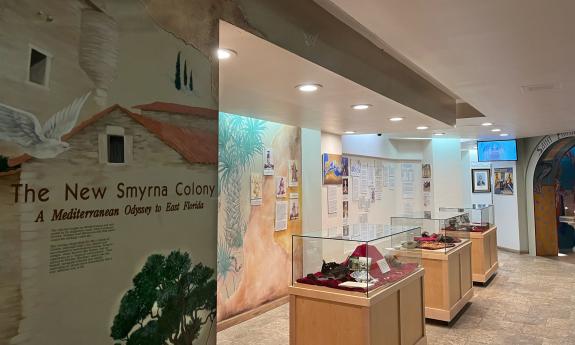 A museum exhibit at St. Photios National Shrine. A row of glass cases displays artifacts on red velvet. Murals and informational panels line the walls