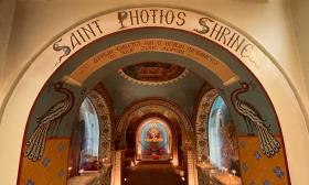 An arched entranceway to the shrine at St. Photios, which reads 'St. Photios Shrine' in painted letters. A row of arches and murals continues below