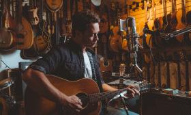 Amos Lee, sitting in a luthier's workshop, holding a guitar, and playing