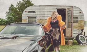 Elle King, wearing an orange coat with feathered collar, standing next to a car and in front of a broken-down camper