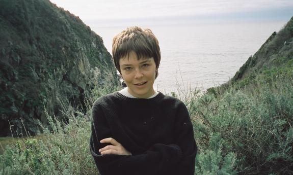 Maggie Rogers standing in a valley with the ocean at her back.