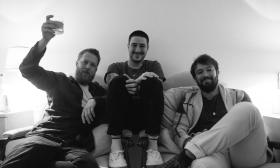 The three members of Mumford and Sons, sitting on a sofa, smiling, and raising a glass