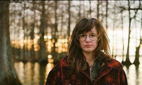 S.G. Goodman - a white woman with tousled hair, trendy glasses, and a flannel jacket, viewable from the shoulders down - in a cypress swamp at sunset