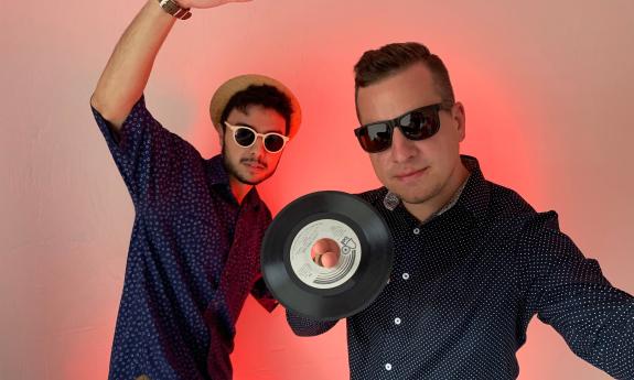 Andre Martins and Ian Opalilnski, standing against an orange background and holding a 45 record