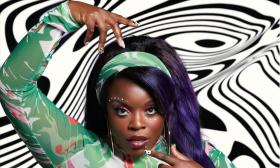 Yola wearing a 70s-inspired dress and headband, standing in front of a black and white-patterned wall