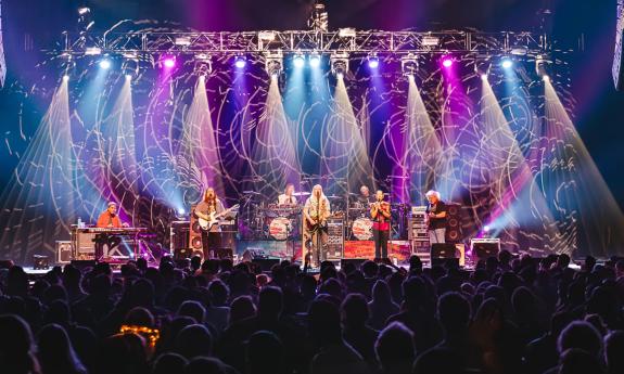 Band members of the Dark Orchestra performs live on a stage with colorful lights. 