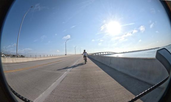 A wide, fish-eye photo of a woman biking across Vilano Bridge after sunrise