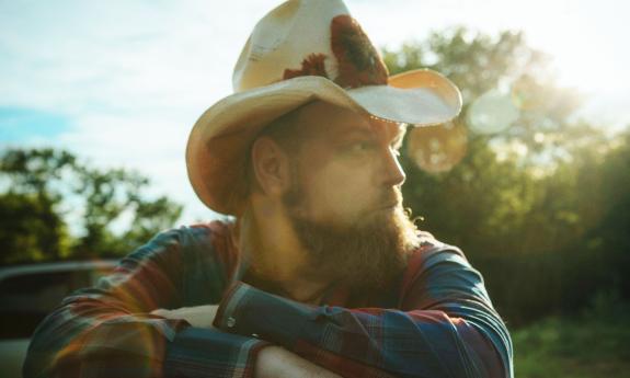 Songwriter Bobby Pinson in cowboy hat leaning on truck, looking sideways.