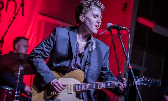 Gabe Stillman sings on a stage with a red backdrop. 