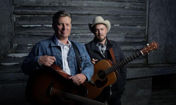 The Gibson Brothers pose with their guitars in front of a wooden background. 