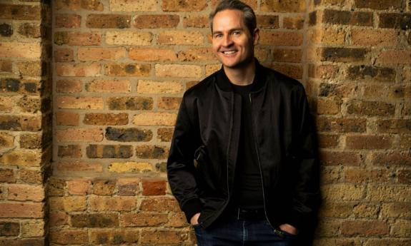 Pat McGann smiles and poses in a leather jacket in front of a brown brick wall. 