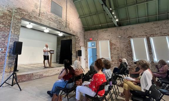 One man stands on stage at a podium, in a small, brick-walled venue, reading poetry