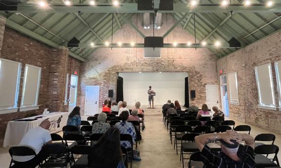 One person standing on stage facing audience, taken from the back of the brick-walled venue