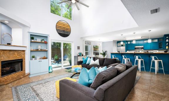 The open-plan living area at Casa Del Sol, has a soaring ceiling, a fireplace, sofas, and a teal kitchen with bar stools