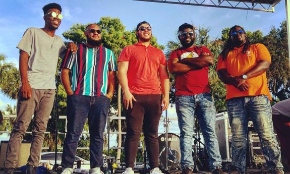 The five members of BlueMoon, standing on an outdoor stage on a sunny day