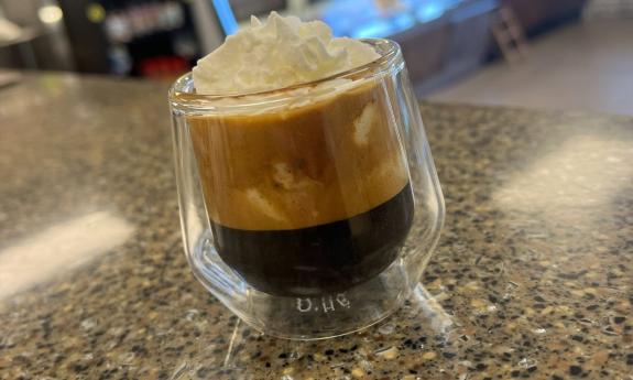 A macchiato in a clear glass on a speckled countertop, topped with a dollop of whipped cream