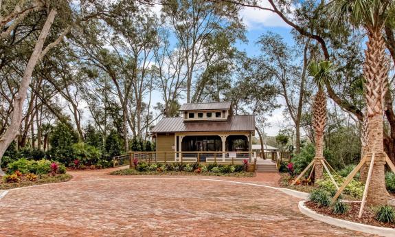 A pavilion with a metal roof and wrap-around porch surrounded by mature trees and tropical foliage, fronted by a red brick pathway