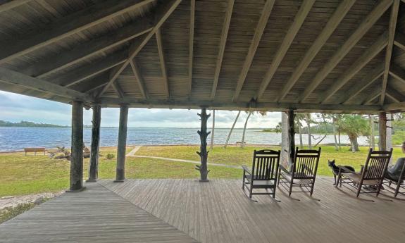 A man sits on a rocking chair at the lodge of Princess Place Preserve, facing Pellicer Creek. His dog looks at the camera.