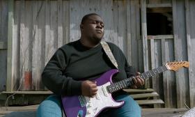 Christone "Kingfish" Ingram, with a purple electric guitar, on steps near an old building