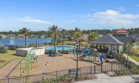 Oxford Estates' gated playground with wood chip ground covering, next to a swimming pool and a lake surrounded by residential houses and palm trees