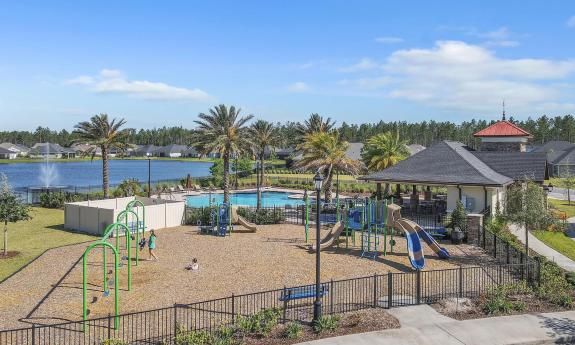 Oxford Estates' gated playground with wood chip ground covering, next to a swimming pool and a lake surrounded by residential houses and palm trees