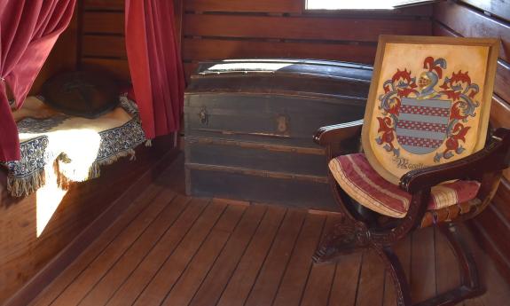 An officer's cabin on the Nao Trinidad is furnished with a trunk, comfortable chair, and bed