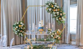 A wedding cake, with white and blue frosting, on a table festooned with flowers
