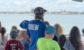 A crewmember of Florida Water Warriors stands at the stern of the boat, surrounded by children in the program