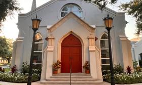 Front entrance of Trinity Parish featuring a Gothic style red door and two lanterns, with lush greenery flanking the steps